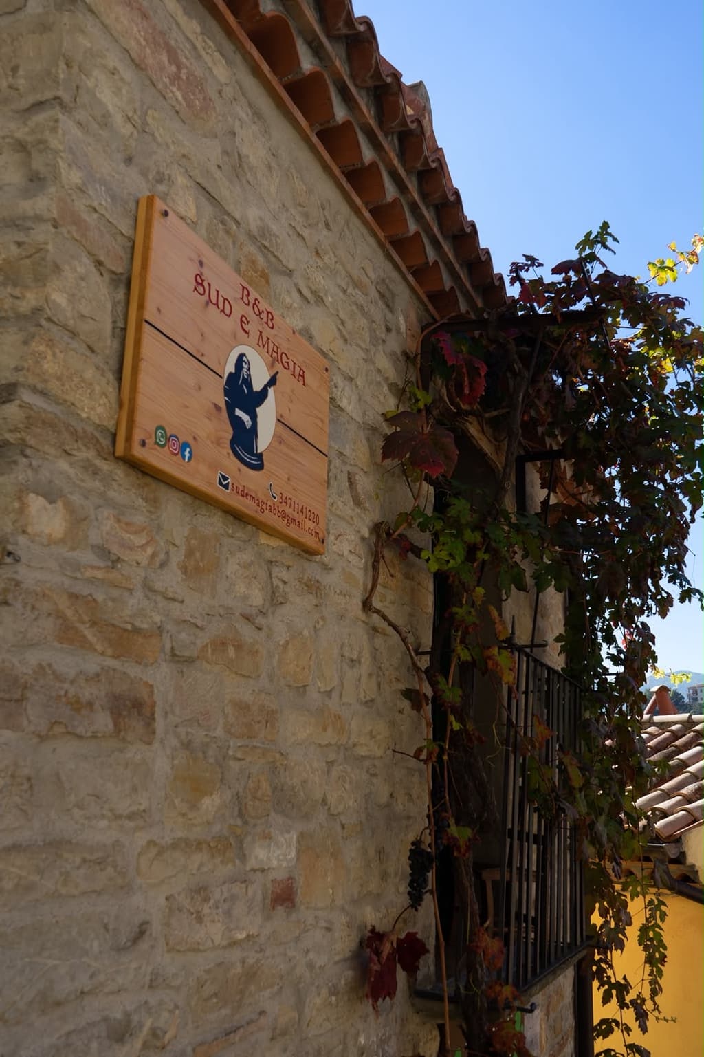 La targa in legno del B&B sul muro in pietra di una casa del centro storico, con la vite rossa sotto il cielo azzurro.